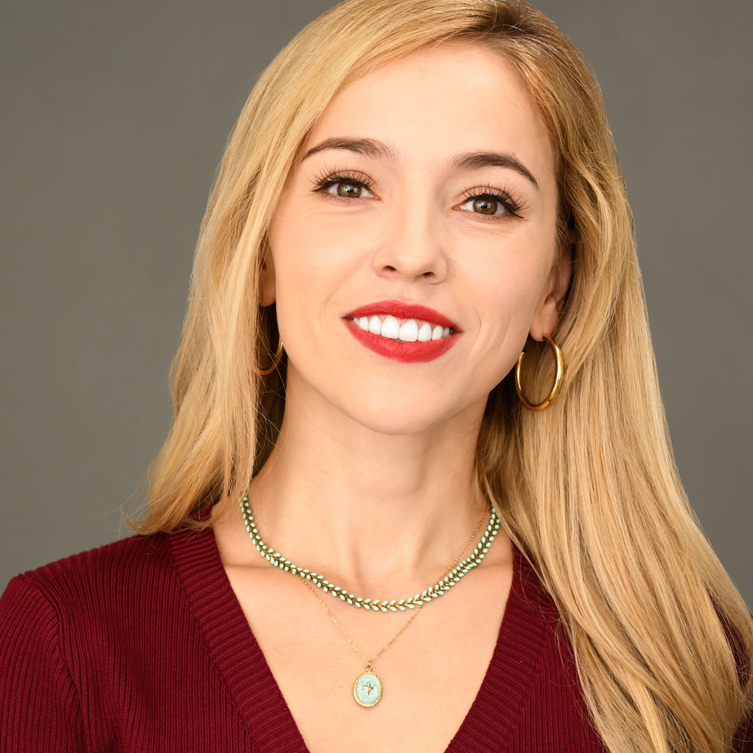 Woman with blonde hair and red lipstick wearing a maroon sweater and high-quality fair-trade gold hoop earrings against a gray background, like Kendra Scott