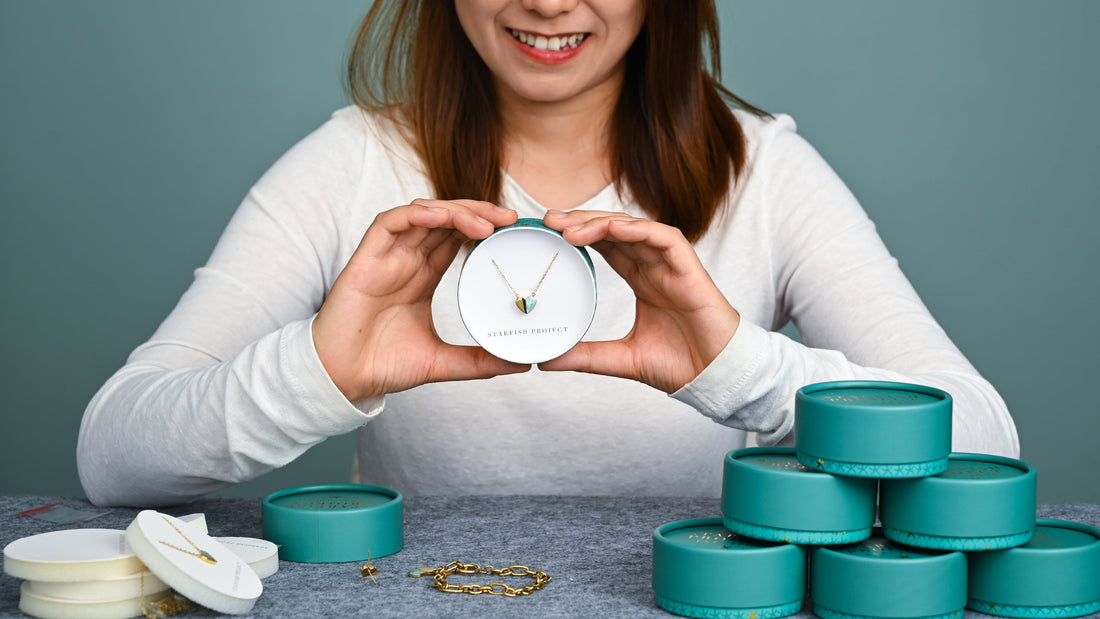 artisan holding a heart shaped fair trade necklace in a round giftable jewelry box.